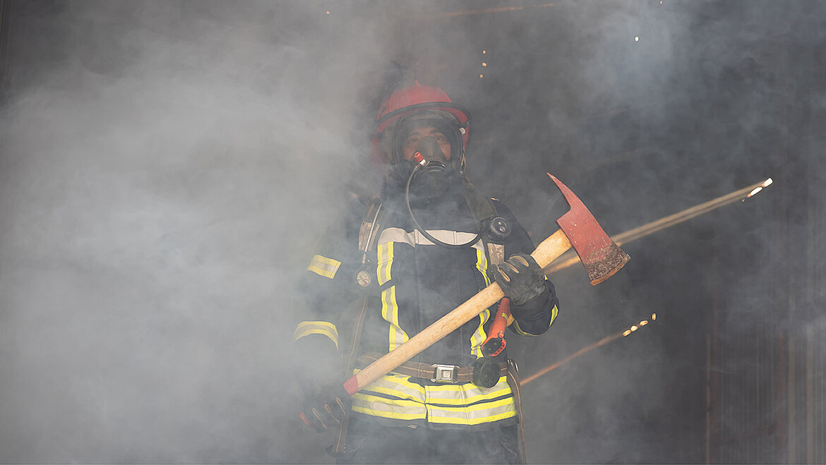 Feuerwehrmann in Schutzkleidung mit Axt, umgeben von dichtem Rauch bei einem Einsatz.