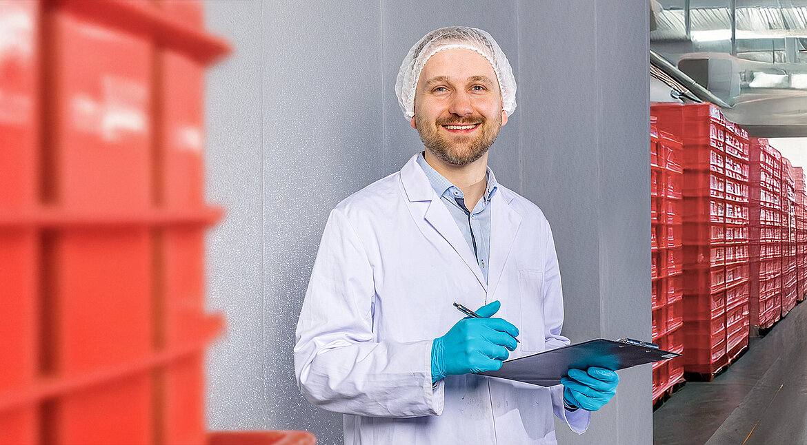 Mitarbeiter in Hygienekleidung mit Klemmbrett steht zwischen roten Transportkisten.