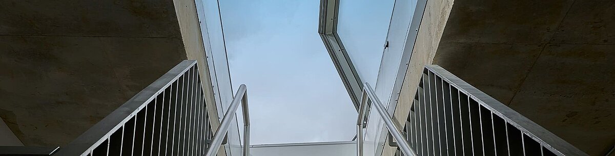Blick von unten auf eine Betontreppe mit Metallgeländern, die zu einem geöffneten LAMILUX Flachdach Ausstieg führt.