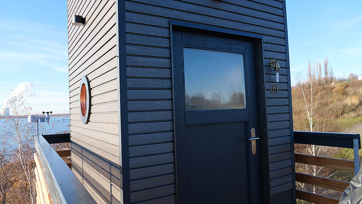 Eingang des DIY-Leuchtturms mit dunkelblauer Holzverkleidung und Bullaugenfenster, umgeben von Natur
