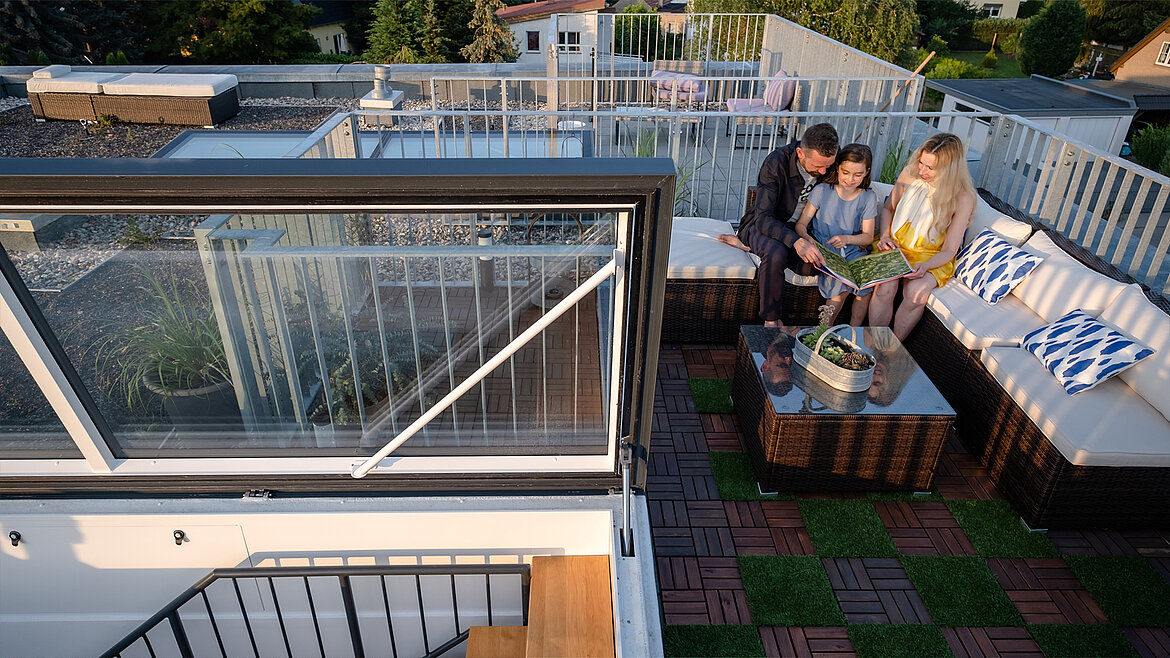 Familie sitzt auf einer Dachterrasse mit Lounge-Möbeln und schaut gemeinsam ein Buch an.
