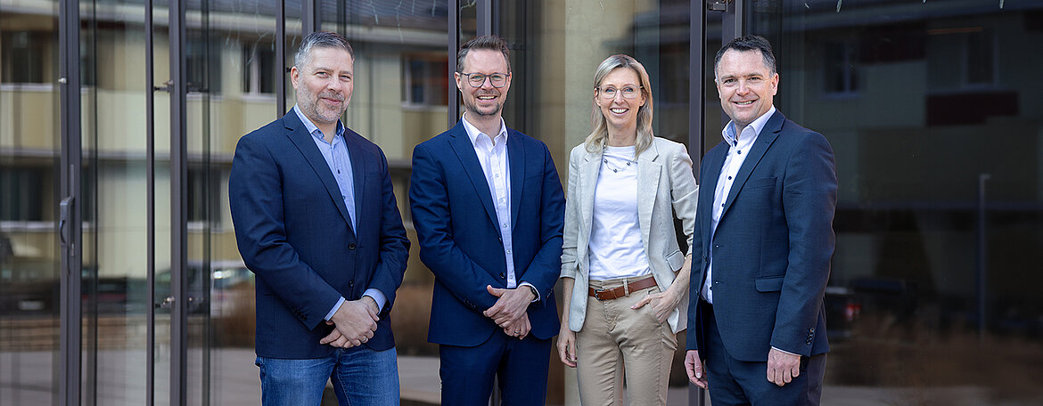 Vier Personen in professioneller Kleidung stehen draußen vor einem Gebäude und lächeln in die Kamera