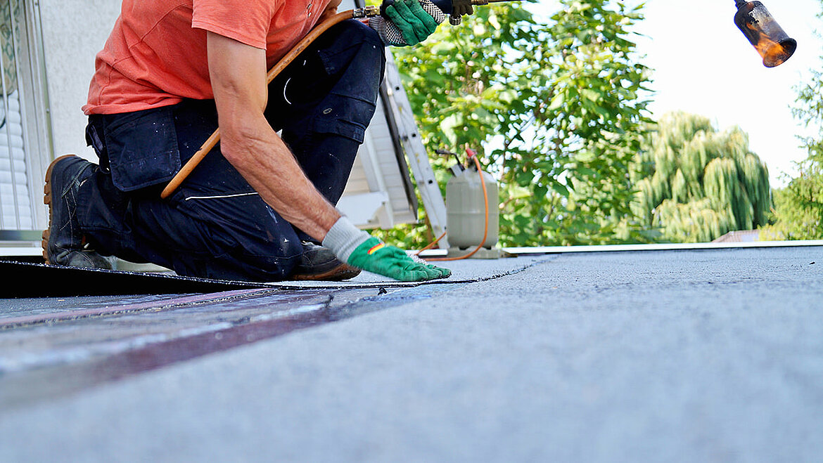 Handwerker kniet auf Flachdach und bringt Abdichtungsmaterial an