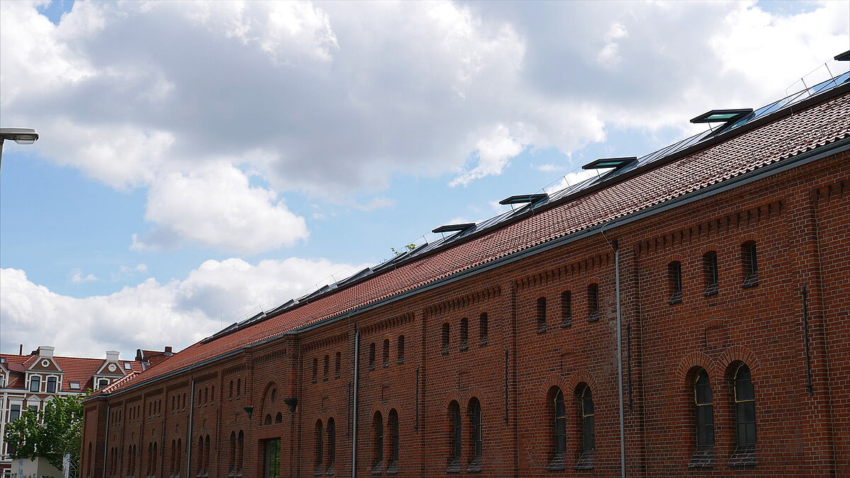 Rotes Backsteingebäude der Leibniz-Universität Hannover mit modernem Glasdach und geöffneten Oberlichtern unter bewölktem Himmel.