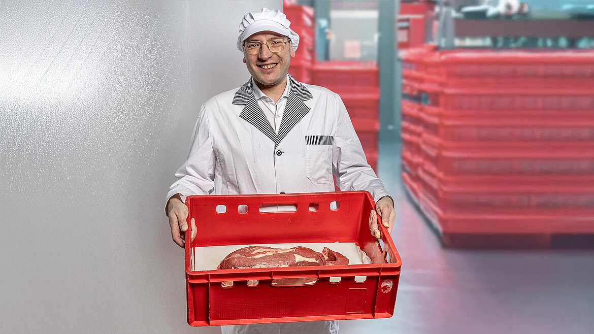 Metzger in weißer Arbeitskleidung hält eine rote Kiste mit Fleischwaren.