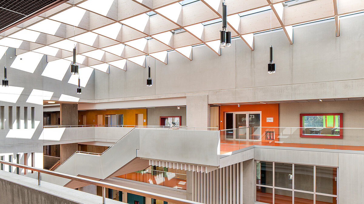 Innenraum des Willibald-Gluck-Gymnasiums in Neumarkt mit Glasdach von LAMILUX, moderner Betonarchitektur und farbigen Akzenten.