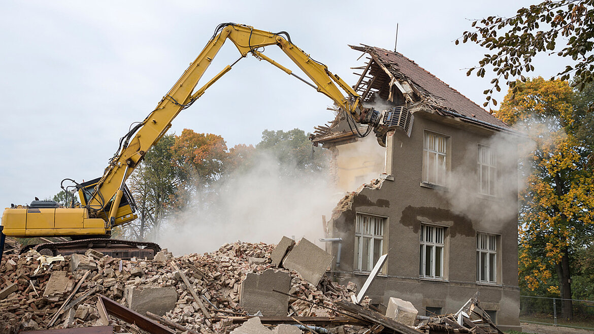 Ein Bagger steht auf einem Haufen Bauschutt und reißt ein graues Gebäude ab.