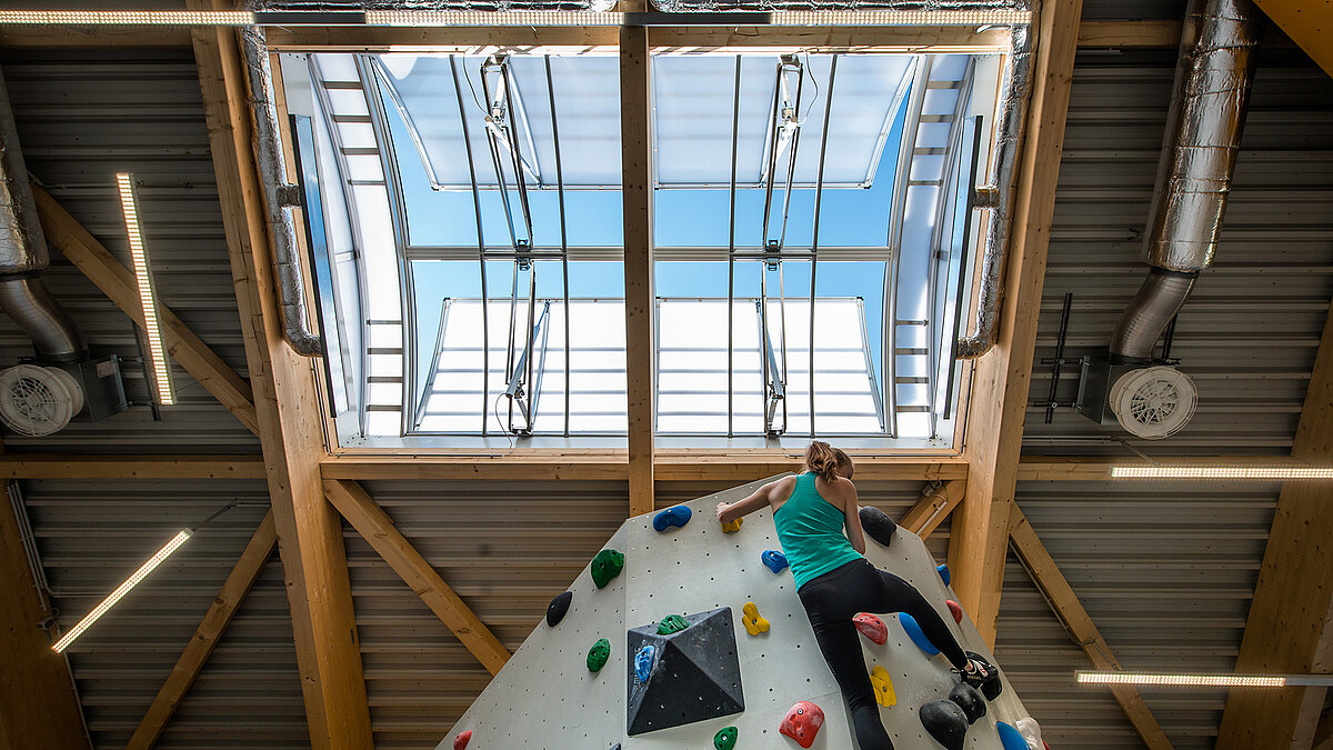 Kletterzentrum OWL mit Kletterwand und geöffneter Lichtkuppel, durch die Tageslicht in die Halle fällt. Eine Frau klettert die bunte Wand hoch.