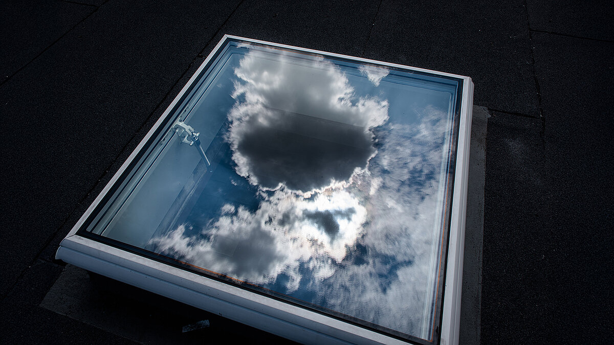Geschlossenes Flachdach-Fenster auf dem Dach des Caritas-Gebäudes in Hagen, mit Spiegelung von Wolken im Glas.