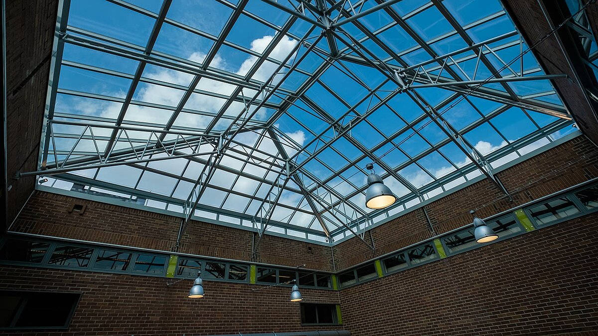 Glasdach des Dante-Gymnasiums mit Stahlträgern und modernen Hängeleuchten, Blick auf blauen Himmel.