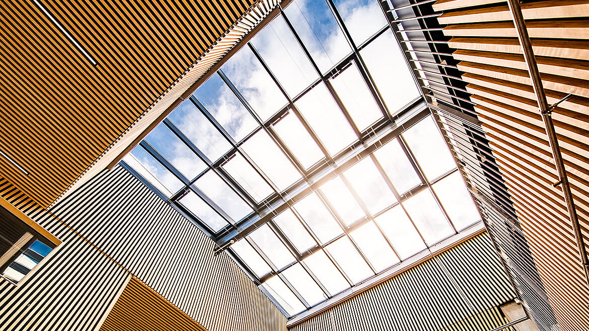 Architektonisch auffälliges Glasdach mit wellenförmigen Holzelementen und Sonnenlicht.