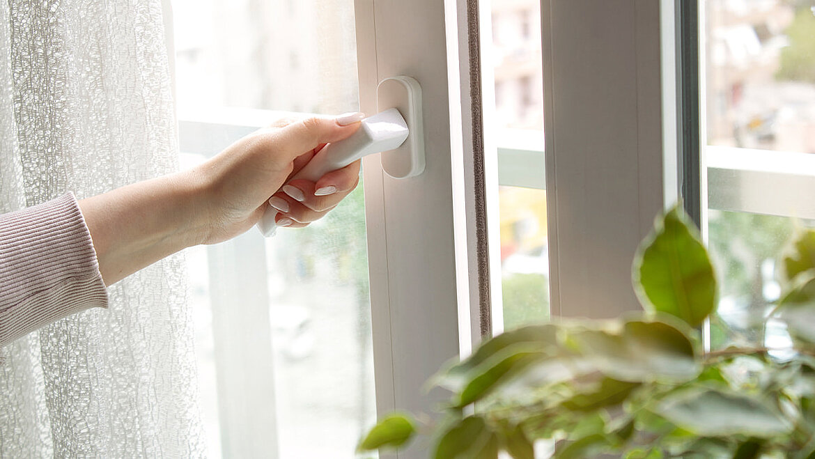 Hand öffnet ein Fenster zum Lüften, um Feuchtigkeit und Kondenswasser im Raum zu reduzieren.