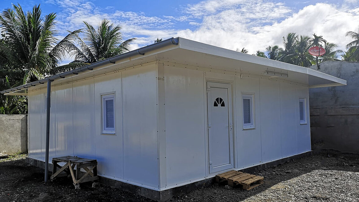 Weißes kleines Haus mit Flachdach und kleinen Fenstern, umgeben von Bäumen