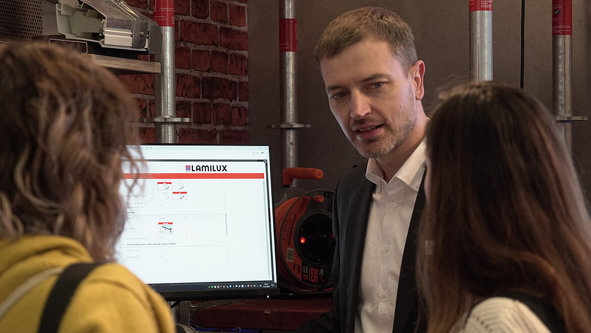 Bauingenieur Carsten Ficker erklärt LAMILUX Produktkonfigurator Carsten Ficker Leiter LAMILUX Academy auf digitalBAU mit zwei Messestandsbesucherinnen | insights by LAMILUX