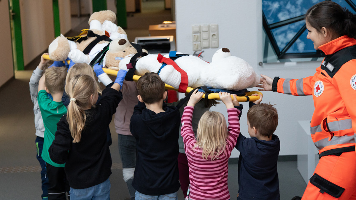 Kinder tragen gemeinsam einen großen Stoffbären auf einer Trage, betreut von einer Person in orangefarbener DRK-Uniform während eines Kinderbetreuungsprogramms am Buß- und Bettag bei Lamilux.